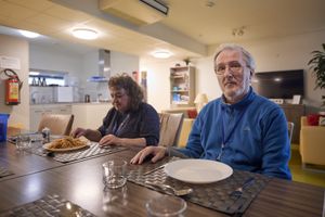 Een man met blauwe trui, grijs haar en een bril en een vrouw met grijze trui zitten aan een gedekte tafel. Op de achtergrond zie je een woonkamer met keuken.