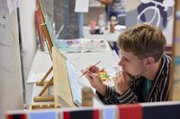 Een jongeman met gestreept overhemd kijkt geconcentreerd naar een schildersdoek.