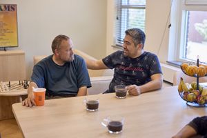 Twee mannen zitten aan een houten tafel met een kop koffie voor zich. Ze kijken elkaar aan en zijn in gesprek.