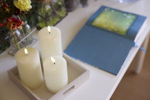 Brandende kaarsen op een dienblad naast een open boek met een blauwe kaft en een groene bladzijde, dat op een witte tafel ligt naast een boeket bloemen.