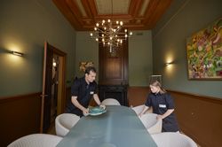 Een man en een vrouw met zwarte kleding zetten borden op een lange tafel met nette stoelen eromheen. Ze staan in een antiek uitziende zaal met aan het plafond een kroonluchter en aan de muur een kleurrijk schilderij.