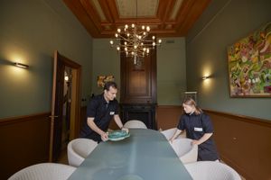 Een man en een vrouw met zwarte kleding zetten borden op een lange tafel met nette stoelen eromheen. Ze staan in een antiek uitziende zaal met aan het plafond een kroonluchter en aan de muur een kleurrijk schilderij.
