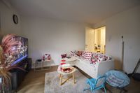 Kleine woonkamer met een witte L-vormige bank met kussens met hartjesmotief, een tv op meubel, een ronde salontafel en een blauwe kinderstoel.