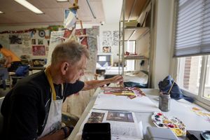 Een man met een schort zit in een ruimte vol schilderijen en tekent aan tafel zelf een schilderij met allerlei verschillende mensen.