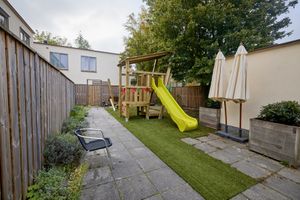 Omheinde achtertuin met een houten speeltoestel en een felgele glijbaan, kunstgras en ingeklapte parasols.