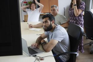 Een man met donker haar en een baard zit achter een computer. Achter hem kijken drie mensen mee naar het scherm.