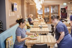Een restaurantmedewerkster met donkerblauw shirt en schort serveert een salade aan een vrouw aan een houten tafel.
