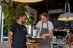 Twee mannen staan in een café met grote plant en een bar. De man met blond haar en een schort zet een kop koffie op een dienblad in de hand van de man met het zwarte shirt.