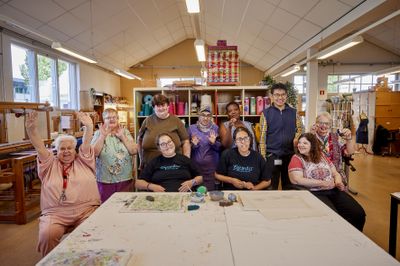 Een diverse groep van tien mensen poseert lachend voor de camera in een grote lichte ruimte vol weefgetouwen en andere creatieve spullen.