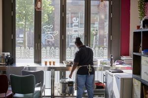 Je ziet een vrouw met een zwarte knot vanaf haar rug. Ze roert in een metalen schaal op een metalen tafelblad aan een groot raam aan de straat met daarop stickers van Amsterdamse grachtenpandjes.