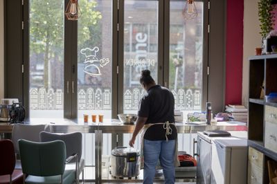 Je ziet een vrouw met een zwarte knot vanaf haar rug. Ze roert in een metalen schaal op een metalen tafelblad aan een groot raam aan de straat met daarop stickers van Amsterdamse grachtenpandjes.