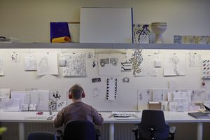 Een man met rode koptelefoon zit aan een groot bureau. De wand hangt vol met schetstekeningen.