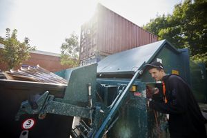 Een jongen met een zwarte jas en pet trekt aan een hendel van een grote blauwe afvalverwerkingsmachine met daaraan vast een container vol met karton.