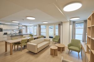 Moderne open woonruimte met keuken, eethoek en zithoek, voorzien van licht houten meubilair, groene stoelen en grote ramen.