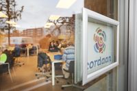 Close up door een raam naar binnen. Op het raam is het logo van Cordaan te zien en de reflectie van de bomen en gebouwen in de omgeving. Door het raam is een werkplek te zien met tafels en stoelen, waar mensen aan zitten en sorteerwerk doen.