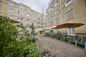 Buitenaanzicht van het gebouw met vijf verdiepingen. Op het terras beneden staan stoelen en parasols, daarachter de ingang met gele deuren.
