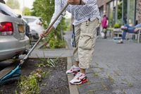 Een man met sportieve Nike Air Max-schoenen aan harkt de  tuin die grenst aan een parkeerplaats.