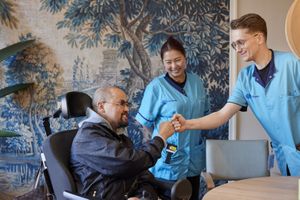 Naast een man in een rolstoel staan twee jonge zorgmedewerkers in blauwe uniformen. Een van de twee zorgmedewerkers geeft de man een boks.