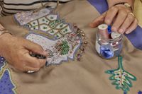 Een paar handen van een ouder persoon hangen boven een handwerkstuk. De linkerhand rust op een glazen potje met gekleurde klosjes garen erin, de rechterhand legt een kraaltje neer. ed voor haar zijn twee zeepaardjes genaaid, een blauwe en een crèmekleurige, met pailletjes erop