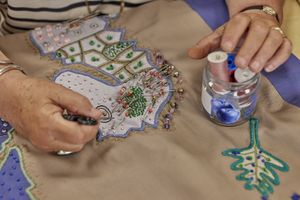 Een paar handen van een ouder persoon hangen boven een handwerkstuk. De linkerhand rust op een glazen potje met gekleurde klosjes garen erin, de rechterhand legt een kraaltje neer. ed voor haar zijn twee zeepaardjes genaaid, een blauwe en een crèmekleurige, met pailletjes erop