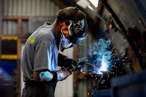 Iemand in een grijs shirt is aan het lassen en draagt een las helm en leren handschoenen.