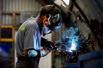 Iemand in een grijs shirt is aan het lassen en draagt een las helm en leren handschoenen.
