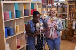 Twee vrouwen staan hard lachend voor een grote kast vol met garen en houden allebei een konijnenknuffel vast, gemaakt van verschillende stoffen met gekleurde prints.