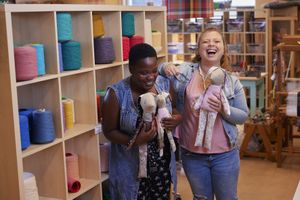 Twee vrouwen staan hard lachend voor een grote kast vol met garen en houden allebei een konijnenknuffel vast, gemaakt van verschillende stoffen met gekleurde prints.
