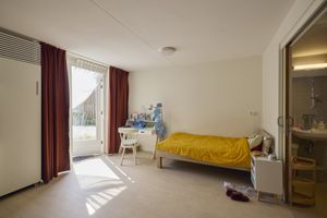 Een lichte kamer met een bed bedekt met een gele deken, een bureau met een stoel en een open deur naar buiten. Rechts is een kleine badkamer zichtbaar.