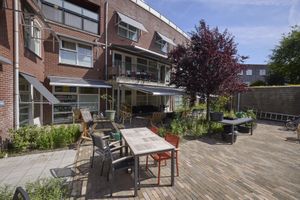 Een ruim terras met tuintafels en -stoelen, omgeven door een bakstenen gebouw, groen en een kleine boom onder een helderblauwe hemel.