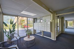 De lobby van het appartementencomplex heeft brievenbussen, twee stoelen, een tafel met een plant, grote ramen en een gang.