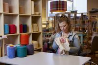 Een vrouw met blonde paardenstaart zit naast een kast met rollen garen te naaien op een borduurring.