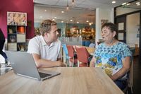Twee personen zitten in gesprek aan een tafel in een algemene ruimte met vrolijke kleuren. Op de achtergrond is een bar te zien. De man links luistert aandachtig en de persoon rechts vertelt.