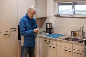 Een man met grijs haar en een bril staat in een keuken. Hij maakt met een theedoek een mes droog.
