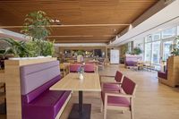Ruim restaurant met houten interieur, paarse zitmeubels, planten en grote ramen die veel natuurlijk licht binnenlaten.