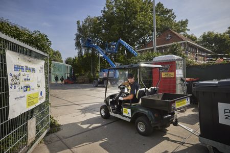 Een man met zwarte outfit en zwarte pet rijdt in een tweepersoons kar met daaraan een container over een industrieterrein. Op de achtergrond staan twee grote, blauwe hoogwerkers.