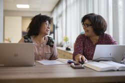 Twee vrouwen zitten in een lichte ruimte naast elkaar aan tafel met laptops voor zich en kijken elkaar lachend aan.