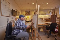 Een man met geruit overhemd en blauwe pet zit in een ruimte vol tekeningen voor een schildersezel te tekenen.