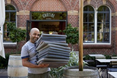 Een kale man staat op een terras van een restaurant en houdt met een blij gezicht een stapel stoelkussens vast.
