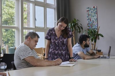 Een vrouw in een jurk kijkt over de schouder van een man mee naar een schrijfblok op tafel.