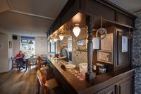 Een ruimte met een grote houten bar in oude stijl met gezellige verlichting en versiering. Achter de bar staat een vrouw met een schort aan.
