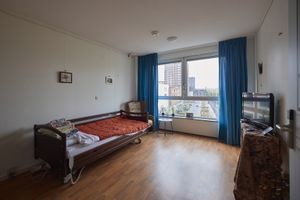 Een kamer met een eenpersoonsbed, schilderijtjes aan de muur, een tv en een groot raam met blauwe gordijnen dat uitzicht biedt op de stad.