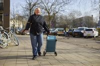 Een blonde dame met zwart koksuniform loopt buiten over de stoep met een boodschappentrolley.