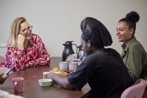 Drie vrouwen zitten samen aan tafel met daarop theekannen en mokken. Ze lachen.