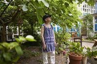Een vrouwelijk deelnemer met pet en een blauw schort aan, kijkt omhoog. Ze neemt eventjes pauze van haar drukke werkzaamheden in de tuin, maar heeft nog wel een snoeischaar in de hand.