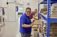 Iemand pakt een platte kartonnen doos uit een stellingkast. Hij kijkt lachend naar de camera. Op de achtergrond werkt iemand aan een tafel. De ruimte is groot. De vloer en muren zijn wit.