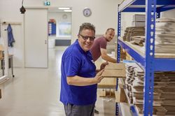 Iemand pakt een platte kartonnen doos uit een stellingkast. Hij kijkt lachend naar de camera. Op de achtergrond werkt iemand aan een tafel. De ruimte is groot. De vloer en muren zijn wit.