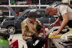 Twee mannen werken aan een spatscherm van een auto. De ene man heeft een hamer in zijn handen en de andere een lasapparaat. Op de achtergrond staat een auto met de motorklep open.