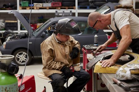 Twee mannen werken aan een spatscherm van een auto. De ene man heeft een hamer in zijn handen en de andere een lasapparaat. Op de achtergrond staat een auto met de motorklep open.
