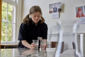 Vrouw met zwarte blouse en twee staartjes zet glazen op tafel voor een raam.